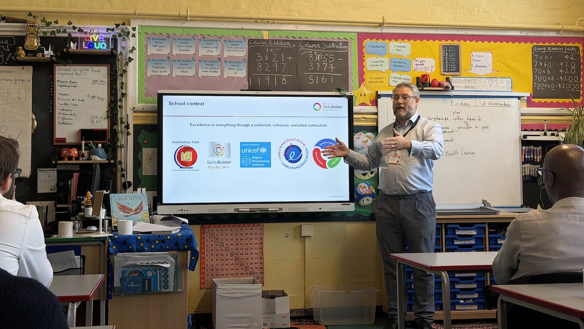 A man delivers a presentation about Shaftesbury Park school, the slide shows logos of several awards the school holds, as well as the school's vision.